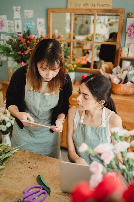 Inside a brightly lit floral shop, two women are engaged in a flower arranging activity. One woman, standing and wearing a black cardigan over a light grey apron, is showing a tablet to the seated wom