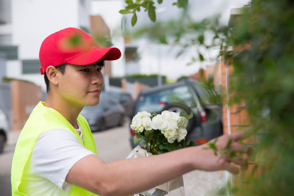A delivery person wearing a red cap and a high visibility yellow vest is placing a bouquet of fresh white roses and greenery against a brick wall outside a residential area. The bouquet is wrapped in 