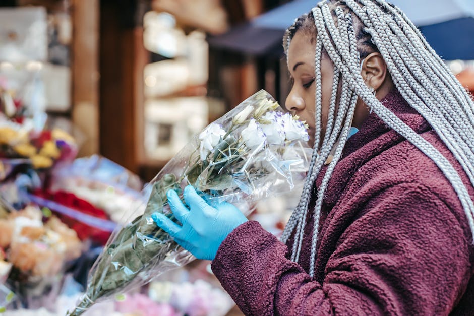 A woman with long, braided, silver-colored hair wearing a maroon fleece jacket and light blue gloves is smelling a bouquet of white roses wrapped in clear plastic at an outdoor flower market. The bouq